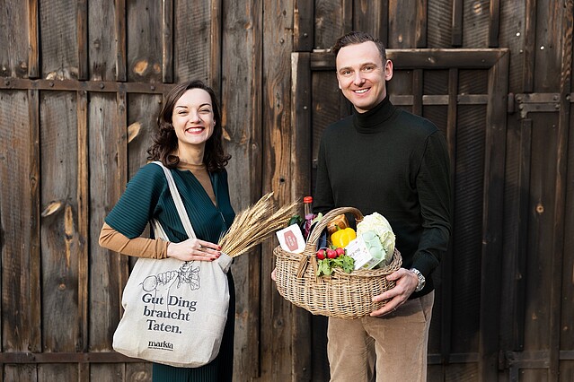 markta: Eine Frau und ein Mann stehen mit einem Getreidesack und einem Obstkorb vor einer Scheunentüre aus Holz. 