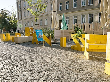 Vor einem Gebäude stehen große, gelbe Holzbuchstaben, die das Wort Urbanize bilden. 