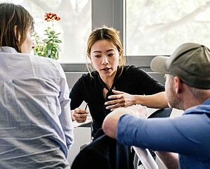 Frau asiatischer Herkunft erzaehlt zwei Maenner mit Ruecken zugedreht hoeren zu