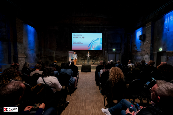 Vortragssaal mit Speakern, Publikum und Bildschirm "Numix Lab"