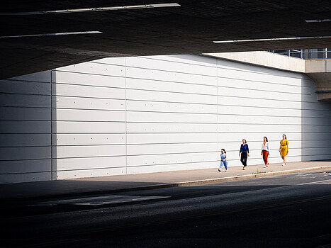 Ein kleines Mädchen und drei Frauen gehen hintereinander an einer weißen Holzwand entlang. 