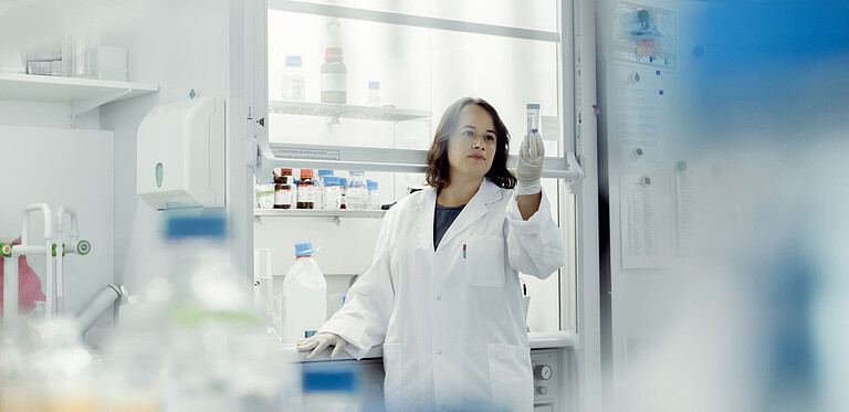 Eine Frau mit Laborkittel und Gummihandschuhen hält ein durchsichtige Flasche in der Hand und betrachtet den Inhalt