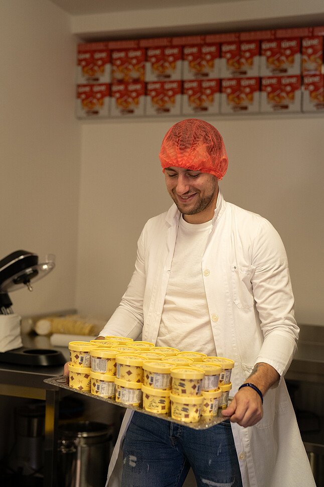 Dajan Srejic trägt ein Haarnetz und einen weißen Kittel und hält ein Tablett mit gelben Eisbechern. Im Hintergrund sind weitere Produkte in Regalen zu sehen.