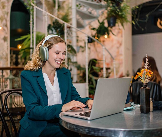 Eine Frau sitzt an einem Tisch und arbeitet an einem Laptop, am Kopf trägt sie ein Headset