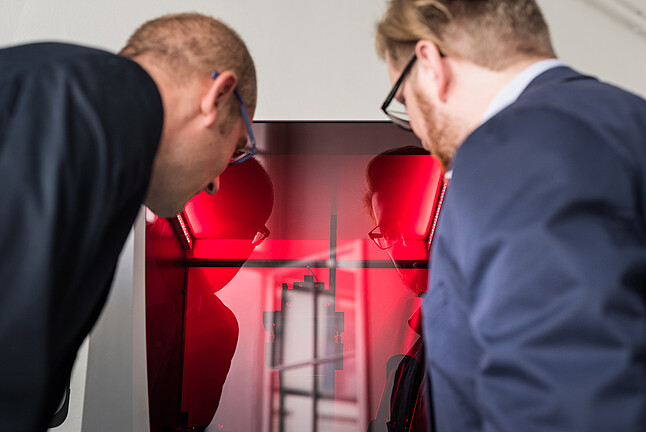 Two men in suits looking into a glass box lit up with a red light