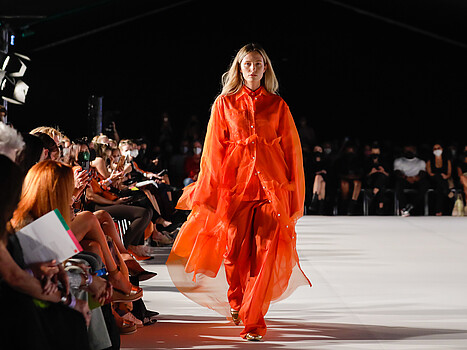 Eine orange gekleidete Frau mit langen blonden Haaren geht über eine Laufsteg