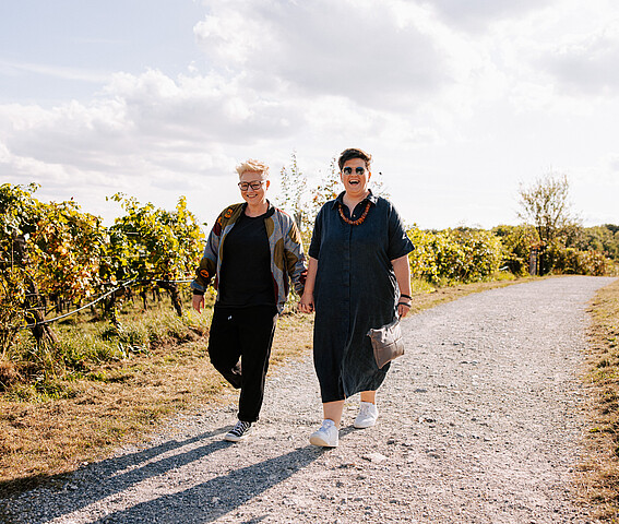 Bistra und Christa: Zwei Frauen gehen Hand in Hand auf einem Schotterweg in den Weinbergen spazieren