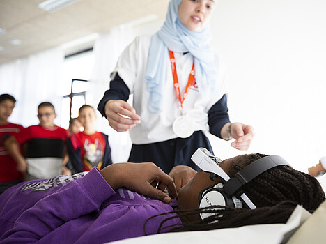 In einem Workshop für Schulklassen steht ein Mädchen gegenüber einem Mädchen, dass eine Datenbrille trägt und auf einem Tisch liegt.