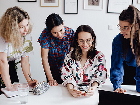 Fashion Revolution Hub: Vier Frauen stehen an einem Tisch und blicken auf ein Handy.