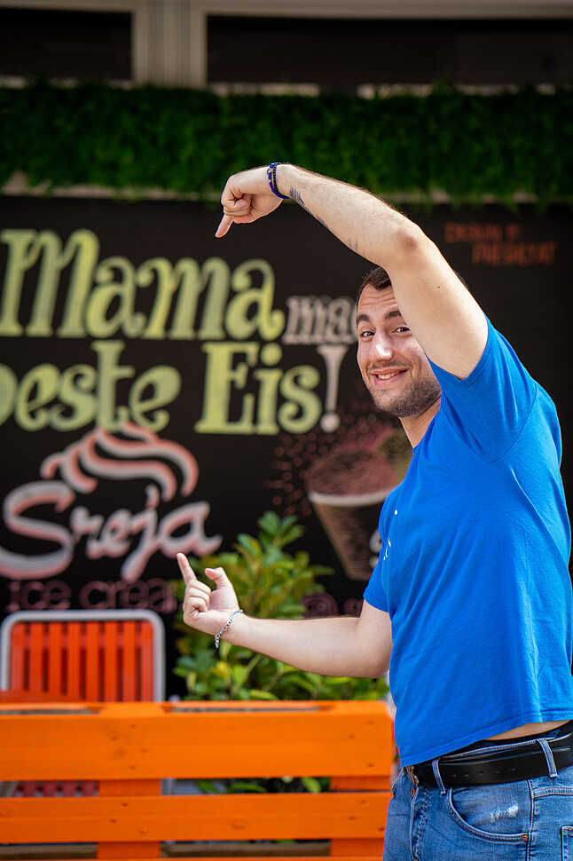 Mann in blauem T-Shirt zeigt lächelnd auf ein Schild mit der Aufschrift „Mama macht das beste Eis!“ im Hintergrund. Es handelt sich um Dajan Srejić, den Gründer von Sreja Ice, der vor seinem Eisladen in Wien posiert.