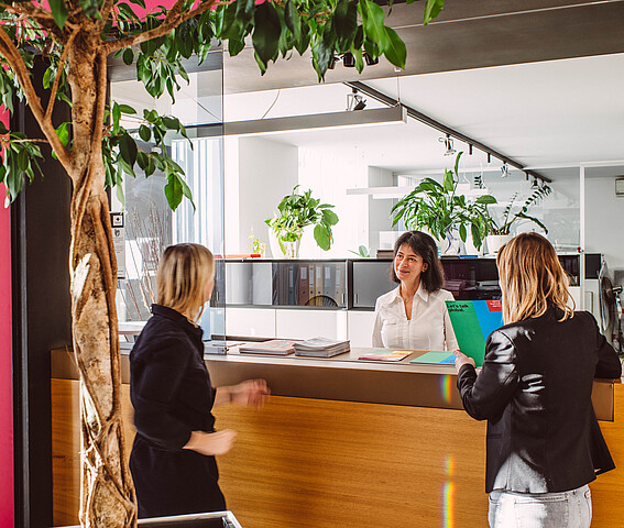 Zwei Frauen werden an einem hellen Empfangsbereich von einer anderen Frau willkommen geheißen. Expat Center: Leben und Arbeiten in Wien.