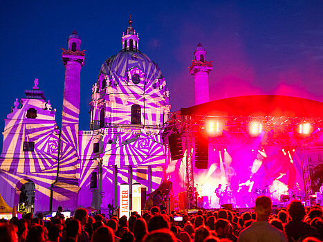 Eine Menschenmenge vor einer pink beleuchteten Bühne, im Hintergrund die Wiener Karlskirche, die ebenfalls bunt angeleuchtet ist.