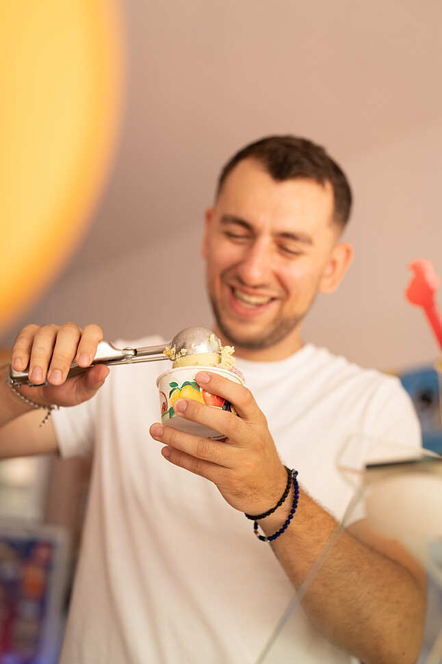 Dajan Srejic, Gründer von Sreja Ice, steht hinter der Theke und lächelt, während er eine Kugel Eis in einen Becher füllt. Er trägt ein weißes T-Shirt und ein Armband, das Eis wird sorgfältig mit einem Eiskugler portioniert.